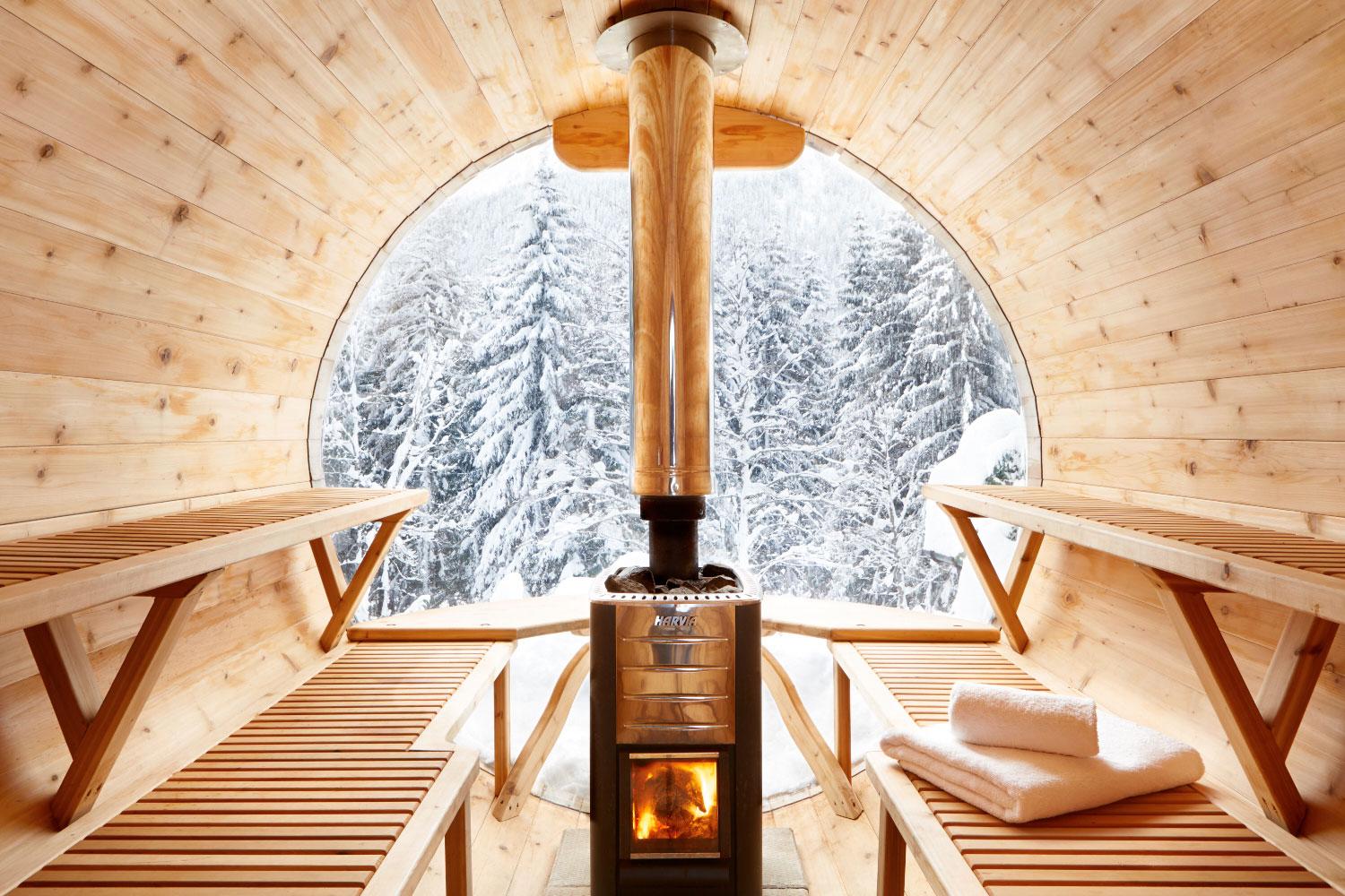 Inside a cedar barrel sauna at sunset, looking out through the round glass end wall to a lake ringed by forested hills. A wood-fired stove burns in the centre between two bench runs, with a cedar bucket and ladle on the bench at right.